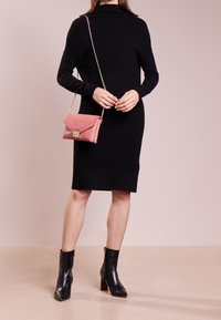 Abito nero in maglia con scollatura alta, abbinato a una borsa a tracolla rosa chiaro con una patta texturizzata e dettagli in metallo dorato, e stivaletti neri alla caviglia.