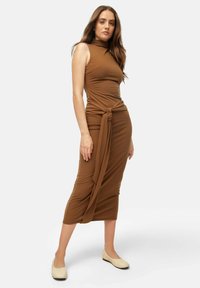 Brown fitted midi dress with a high neck and knotted waist detail. The model wears cream-colored shoes, standing on a plain white background.