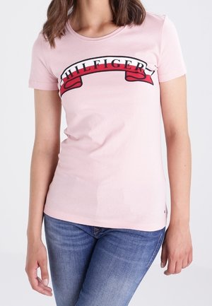 Femme portant un T-shirt rose clair avec "HILFIGER" en lettres noires sur une bande rouge, associé à un jean bleu.