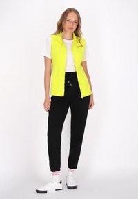 Vesta fără mâneci de culoare galben neon purtată peste un tricou alb, asortată cu pantaloni de trening negri și adidași albi cu accente negre și șosete roz.