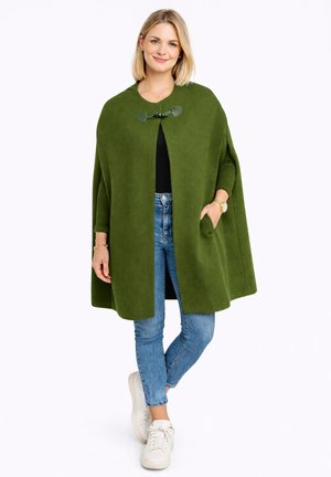 Frau trägt grünen Cape-Mantel über einem schwarzen Oberteil und blauen Jeans, steht mit einer Hand in der Tasche und lächelt.