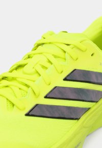 Limettengrüner Sportschuh mit strukturierter Oberfläche, ausgestattet mit schwarzem Drei-Streifen-Design und passenden Schnürsenkeln. Leichtes und schlankes Design.