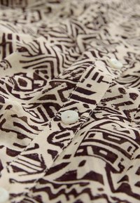 Tejido beige con patrones abstractos en marrón; presenta una tapeta con botones blancos y un detalle de costura visible.