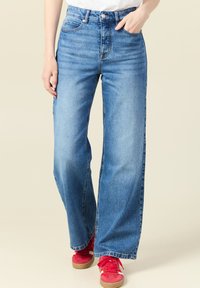 Vaqueros de tiro alto en azul claro con corte de pierna ancha, presentando un clásico diseño de cinco bolsillos y un ligero desgaste. Combinados con zapatillas rojas.