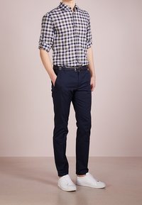 Mężczyzna w podwiniętej koszuli w kratę, granatowych spodniach z czarnym paskiem i białych sneakersach, stojący z jedną ręką w kieszeni na jednolitym tle.