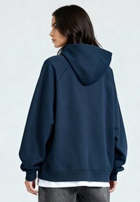 Femme aux cheveux foncés portant un sweat à capuche bleu marine, debout de profil, sur un fond clair uni.