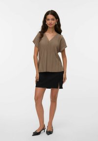 Mujer vestida con una blusa marrón de cuello en V con plisados, falda mini negra con abertura lateral, tacones negros de punta y pendientes de aro, sobre un fondo blanco.