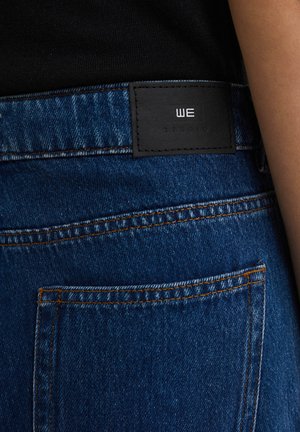 Modré džíny z denimu s čiernou koženou etikettou s embosovaným logom. Šitie v oranžovej farbe, ukazujúce klasický dizajn s piatimi vreckami.
