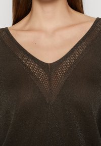 Femme portant un haut en maille scintillante marron foncé avec un col en V orné d'une bordure en mesh à motif.