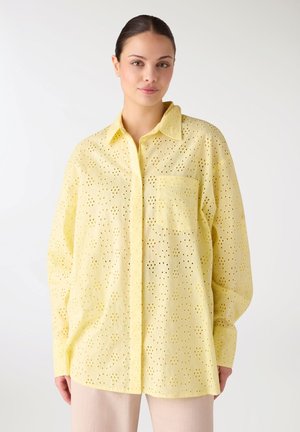 Donna che indossa una camicia gialla larga con motivo a occhielli, con colletto e taschino sul petto, abbinata a pantaloni chiari.