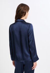 Camisa de satén azul marino con mangas largas, cuello clásico y textura lisa. La parte trasera presenta un diseño sencillo sin adornos.