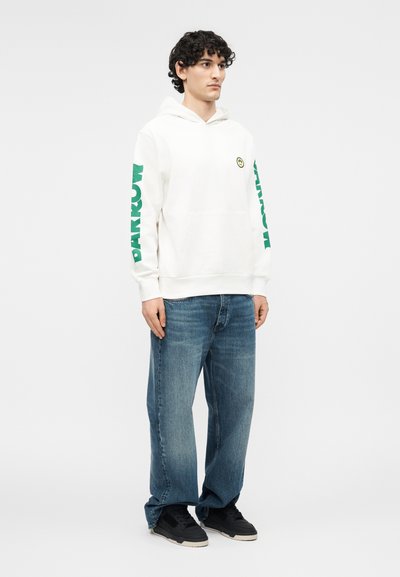 Νεαρός άνδρας με σγουρά μαλλιά, που φοράει λευκή φούτερ με κουκούλα με πράσινο κείμενο "PARKOUR" στα μανίκια, μπλε τζιν και μαύρα αθλητικά παπούτσια, στέκεται προς τα εμπρός.