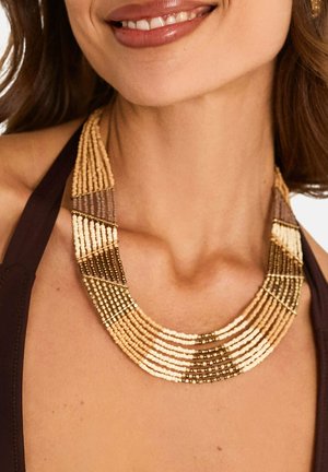 Femme portant un collier à plusieurs rangs de perles aux tons beige, marron et or, assorti à un haut dos nu foncé.