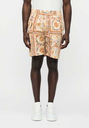 Mann trägt beige Shorts mit Blumen- und Sonnenmuster, weißes T-Shirt und weiße Turnschuhe, steht vor einem schlichten Hintergrund.