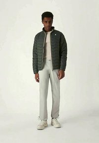 Άνδρας που φοράει ένα σκούρο πράσινο επενδυμένο μπουφάν, λευκό πουκάμισο, ανοιχτό γκρι παντελόνι και λευκά αθλητικά παπούτσια, στέκεται σε απλό φωτεινό φόντο.