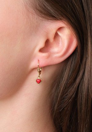 Nahaufnahme eines durchstochenen Ohrs, das einen goldenen Creolenohrring mit einem kleinen, herzförmigen roten Anhänger trägt. Braunes Haar ist im Hintergrund sichtbar.