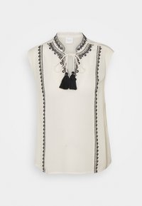 Haut sans manches crème avec détails brodés noirs, doté d'un col à nouer et de deux pompons noirs. Tissu lisse avec une coupe décontractée.