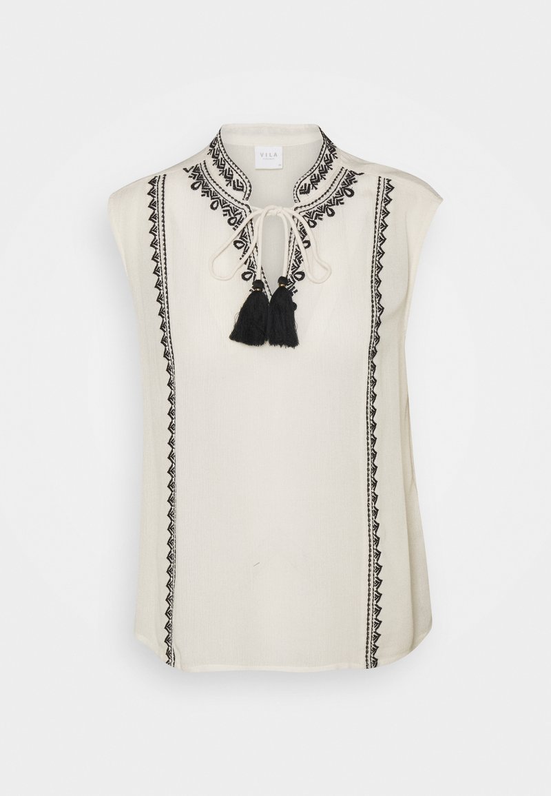Haut sans manches crème avec détails brodés noirs, doté d'un col à nouer et de deux pompons noirs. Tissu lisse avec une coupe décontractée.