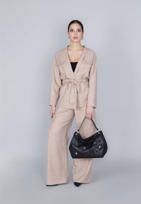 Blazer beige in lino con maniche ampie e cintura, abbinato a pantaloni a gamba larga coordinati. Tiene una borsa nera con accenti in argento e borchie.