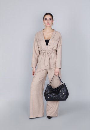 Beige linnen blazer met brede mouwen en riem, gecombineerd met bijpassende wijde broek. Houdt een zwarte handtas met zilveren accenten en studs vast.