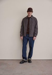 Grå uldjakke med knapper, brun sweater under, blå jeans og sorte sko. Modellen bærer en matchende brun kasket. Enkel, ren design.
