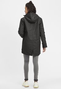 Chaqueta negra impermeable con capucha, diseño de longitud intermedia y ranuras laterales, combinada con leggings grises y zapatillas ligeras.