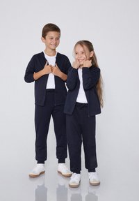 Granatowe kurtki na zamek i spodnie z miękkiego materiału, w zestawieniu z białymi T-shirtami. Dzieci noszą białe trampki z gumową podeszwą; ich uśmiechy są widoczne.