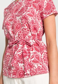 Blommig blus i röd och rosa paisley-mönster med figursydd midja och rosett-accent. Mjuk tyg med korta ärmar och rund halsringning.