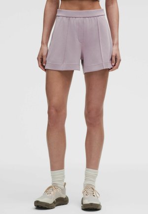 Kvinde iført lyse lilla træningsshorts, hvide ankelsokker og beige løbesko, stående mod en ensfarvet baggrund.