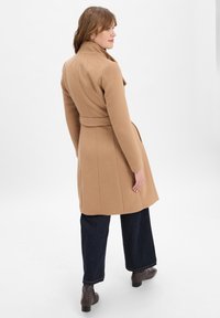 Camel-wolblend jas met een strakke taille, getailleerde pasvorm, lange mouwen en zijzakken. Gecombineerd met brede donkere jeans en bruine enkellaarsjes.