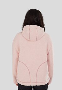 Persoon met lang, donker haar die een lichtroze, gestructureerde, hooded jacket draagt, gezien van achteren tegen een effen grijze achtergrond.