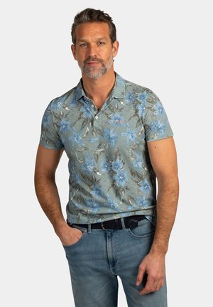 Man met kort haar en baard, gekleed in een groen polohemd met bloemenprint en blauwe spijkerbroek, poserend met één hand in zijn zak tegen een effen achtergrond.