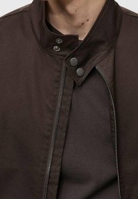 Brauner, leichter Mantel mit hohem Kragen, Druckknöpfen und durchgehendem Reißverschluss. Das Material wirkt glatt mit einer dezenten Struktur.