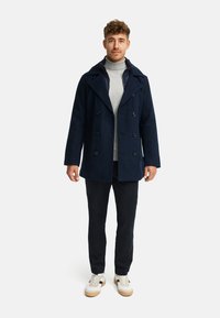 Marineblaßer, doppelt-knöpfiger Mantel mit großen Revers, Knopfakzenten, kombiniert mit einem hellgrauen Rollkragenpullover, dunklen Hosen und weißen Sneakers.