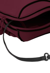 Sac bordeaux avec une finition en tissu lisse, doté d'une fermeture éclair noire et d'une sangle grise réglable attachée à un crochet pivotant. Ouvert pour révéler l'intérieur.