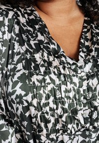 Blouse fleurie présentant des couleurs vertes et blanches, une texture plissée, un design à boutons sur le devant et un décolleté en V profond.