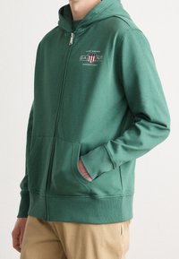 Felpa verde con zip, dotata di cappuccio, tasche frontali e logo ricamato. Texture morbida e vestibilità casual, abbinata a pantaloni beige chiaro.