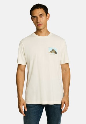 Man met kort krullend haar, die een wit T-shirt met korte mouwen draagt met een kleine bergafbeelding op de linkerborst en een blauwe spijkerbroek.
