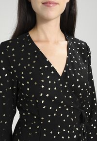 Robe portefeuille noire avec un motif de pois dorés, texture lisse, décolleté en V profond et taille cintrée. Manches longues avec un léger éclat.