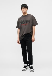 Σκούρο γκρι υπερμεγέθη μπλουζάκι με μαύρο και κόκκινο κείμενο, συνδυασμένο με εφαρμοστά μαύρα τζιν και μαύρα-άσπρα αθλητικά παπούτσια. Μινιμαλιστικός σχεδιασμός.