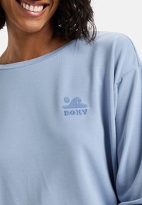 Světle modrá mikina s volným střihem, s kulatým výstřihem a vyšitým logem vlny v odpovídající barvě na hrudi.