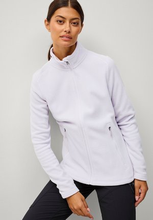 Kobieta o ciemnych włosach związanych w kucyk, ubrana w biały polar z zamkiem i czarne spodnie, pozująca na jednolitym jasnoszarym tle.