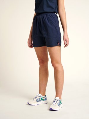 Personne portant un short bleu marine à taille élastique avec poches avant et des baskets blanches avec des détails bleus et verts, debout sur un fond uni.