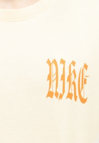 Ύφασμα κρεμ χρώματος με πορτοκαλί γράμματα σε γοτθικό στιλ που σχηματίζουν το "NIKE" στην περιοχή του στήθους ενός ενδύματος.