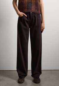 Donkerbruine, losse broek met een hoge taille, voorzien van voorzakken en een metalen knoopsluiting. Gemaakt van zachte, gestructureerde stof.