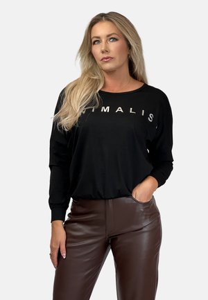 Frau mit langen blonden Haaren trägt ein schwarzes Langarmshirt mit dem Text "MINIMALIS" und braune Lederhosen, Hand in der Tasche, neutraler Hintergrund.