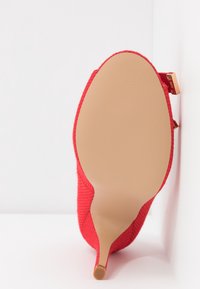 Escarpin rouge à talons hauts avec un dessus texturé, une pointe effilée et une semelle nude. Présente un accent doré sur le talon pour plus de détails.
