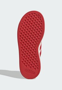 Červená podrážka boty Adidas s hexagonálním dezénem a logem poblíž paty, vrhající stín na světlém pozadí.