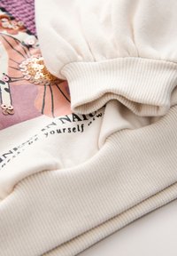 Hanorac de culoare crem cu tiv și manșete cu textură ripă, design floral cu paiete și text imprimat pe partea din față.