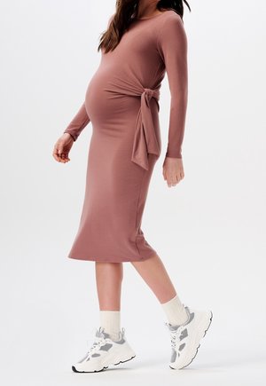 Donna incinta che indossa un vestito midi aderente rosa polveroso con un nodo laterale e sneaker bianche chunky con calzini crema.